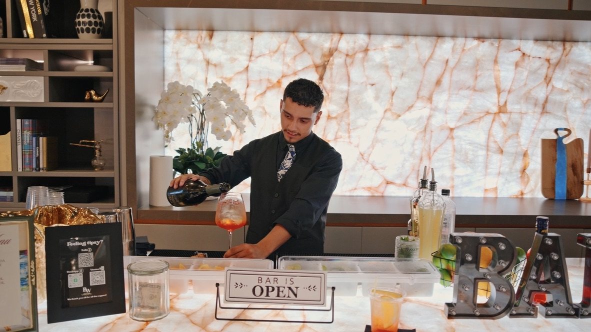Rey pouring a spritz behind a backlit onyx bar with the Bar Is Open sign and marquee BAR letters